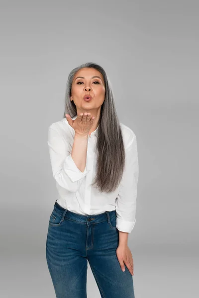 Mediana edad asiático mujer en blanco camisa y azul jeans soplado beso en cámara aislado en gris - foto de stock