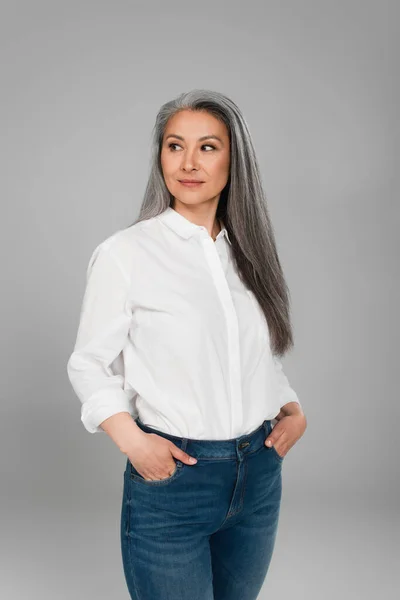 Mujer asiática de mediana edad de pie con las manos en bolsillos de jeans aislados en gris - foto de stock
