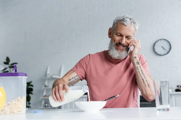 Uomo maturo versando latte in corn flakes e parlando sullo smartphone — Stock Photo