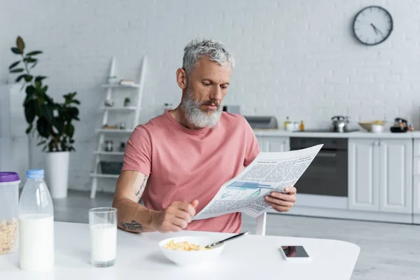 Hombre maduro leyendo periódico cerca de copos de maíz y teléfono inteligente en la cocina - foto de stock