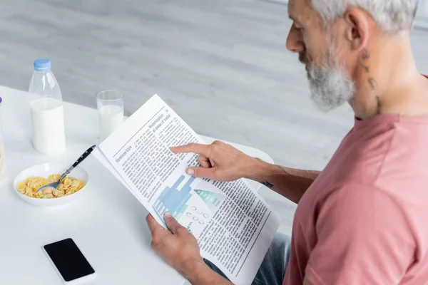 Blurred man reading newspaper near corn flakes, smartphone and milk — Stock Photo