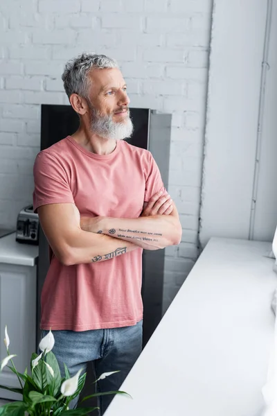 Mature man standing near windowsill and plant in kitchen — Stock Photo