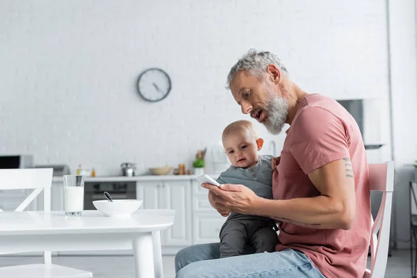 Homme tenant téléphone portable près de tout-petit enfant et petit déjeuner dans la cuisine — Photo de stock