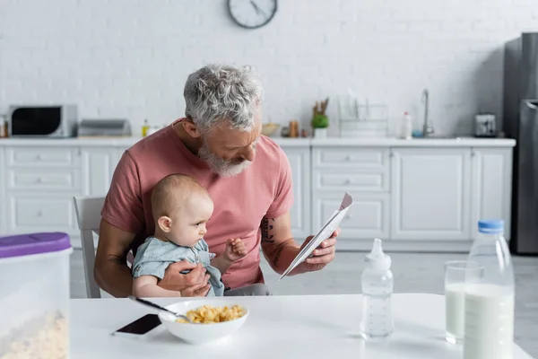 Mature man reading news near baby daughter and breakfast in kitchen — Stock Photo