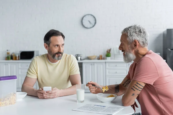 Homosexual man with cornflakes looking at partner using smartphone in kitchen — Stock Photo