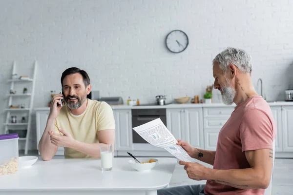 Homosexual hombre hablando en teléfono inteligente cerca de la pareja con el periódico y el desayuno - foto de stock