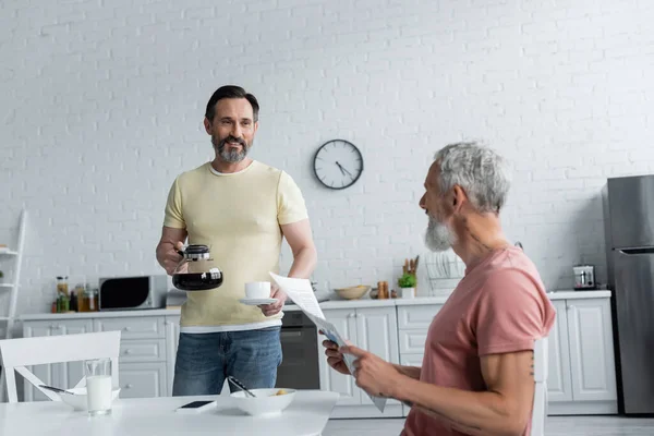 Hombre homosexual sonriente sosteniendo cafetera cerca de la pareja con periódico en la cocina - foto de stock
