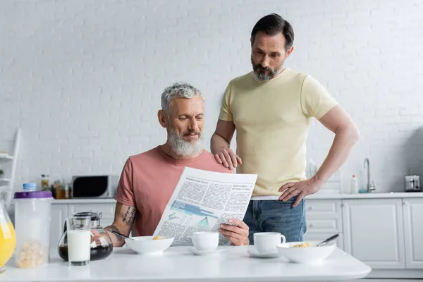 Homosexual couple reading newspaper near breakfast in kitchen — Stock Photo