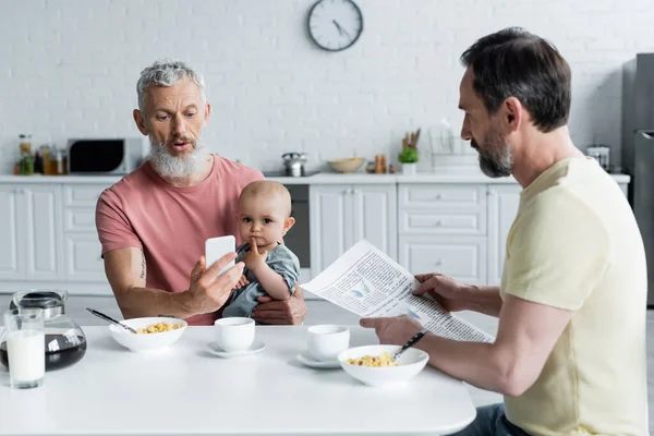 Homosexual man using smartphone near baby daughter and partner during breakfast — Stock Photo