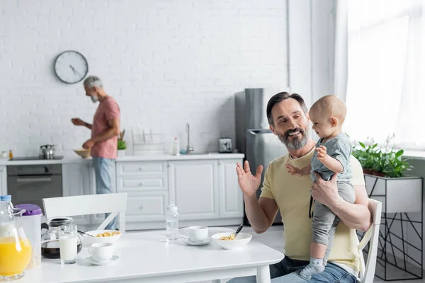 Sorrindo homossexual homem brincando com a filha perto do café da manhã e parceiro turvo — Fotografia de Stock