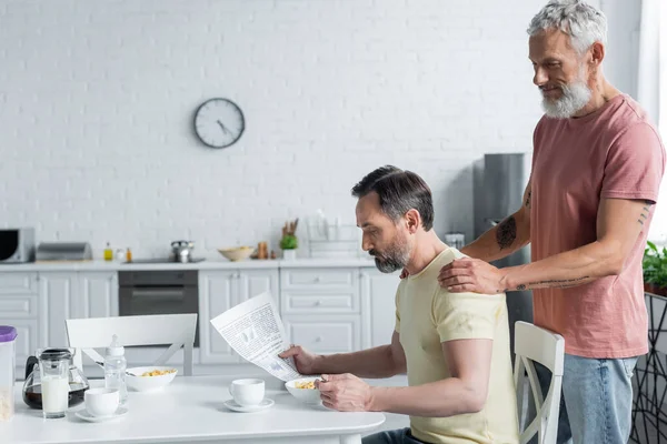 Гомосексуальный мужчина обнимает партнера с газетой на кухне — стоковое фото