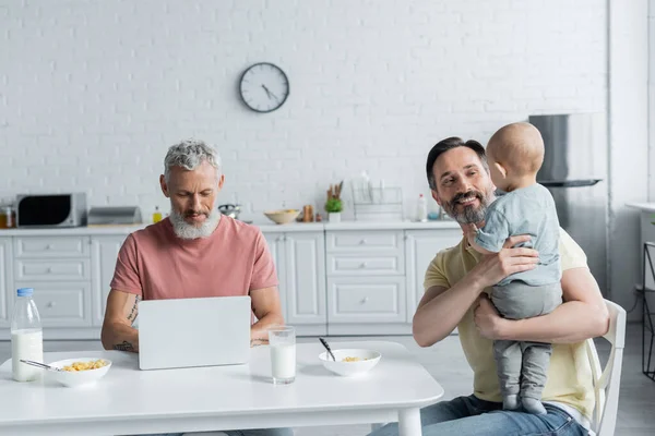 Sonriente hombre homosexual sosteniendo bebé niña cerca de la pareja utilizando el ordenador portátil y el desayuno en la cocina - foto de stock