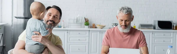 Sorrindo homossexual homem segurando filha perto do parceiro usando laptop na cozinha, banner — Fotografia de Stock