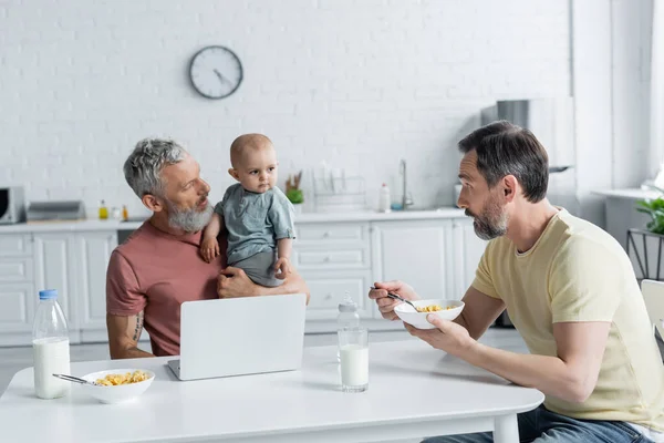 Homosexuelles Paar sieht Baby-Tochter in der Nähe von Laptop und Frühstück — Stockfoto