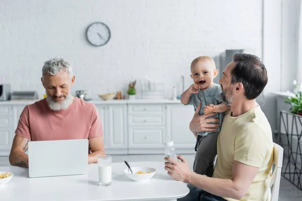 Homosexual man holding daughter and baby bottle near partner using laptop in kitchen — Stock Photo