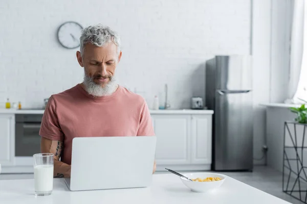 Maduro freelancer utilizando portátil cerca del desayuno en la cocina — Stock Photo