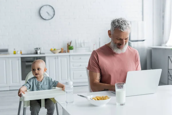 Baby girl sitting on high chair near smiling father using laptop — Stock Photo