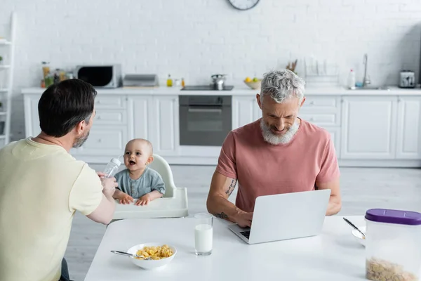 Homosexual man holding bottle near baby and partner with laptop in kitchen — Stock Photo