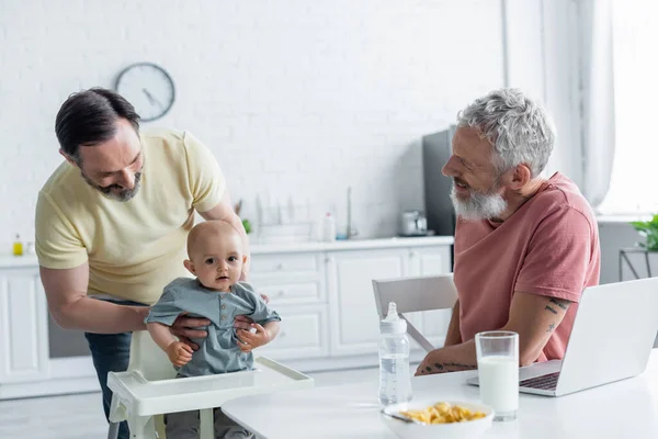 Sonriente pareja homosexual mirando a la hija del bebé cerca de la computadora portátil y el desayuno - foto de stock