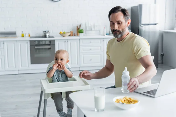 Зрілий чоловік тримає пляшку для немовлят біля ноутбука, сніданок і дочка на високому стільці — стокове фото