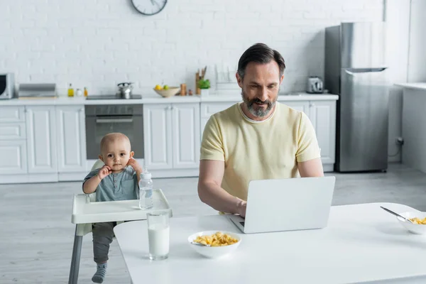 Hombre usando el ordenador portátil cerca del desayuno y el bebé en la silla alta en la cocina - foto de stock