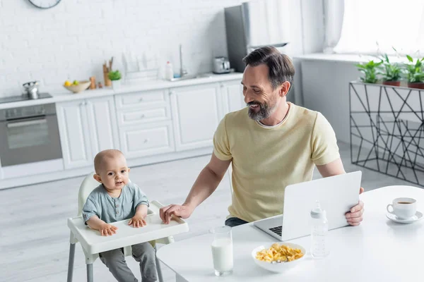 Père souriant avec ordinateur portable regardant enfant sur chaise haute près du petit déjeuner et des boissons — Photo de stock