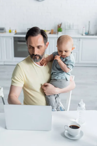 Mature father holding baby daughter and using laptop near coffee in kitchen — Stock Photo