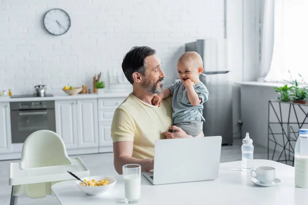 Hombre maduro mirando a la hija del bebé cerca del ordenador portátil y el desayuno - foto de stock