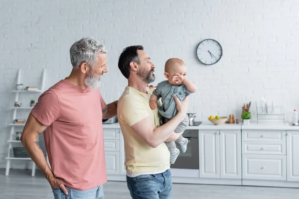 Mismo sexo padres sosteniendo bebé hija en cocina - foto de stock