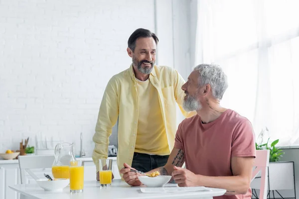 Улыбающийся гомосексуалист стоит рядом с партнером и завтракает на кухне — стоковое фото