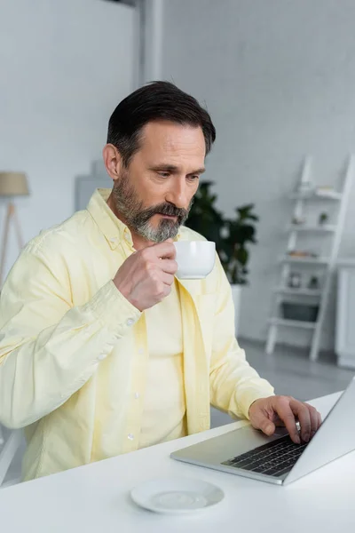 Hombre maduro sosteniendo la taza de café y el uso de ordenador portátil en casa - foto de stock