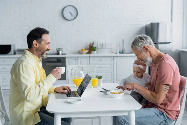 Sonriente hombre homosexual con portátil y café mirando a la pareja con la hija en la cocina - foto de stock