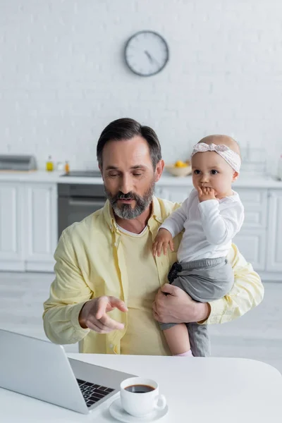 Mature homme pointant vers ordinateur portable tout en tenant bébé fille dans la cuisine — Photo de stock