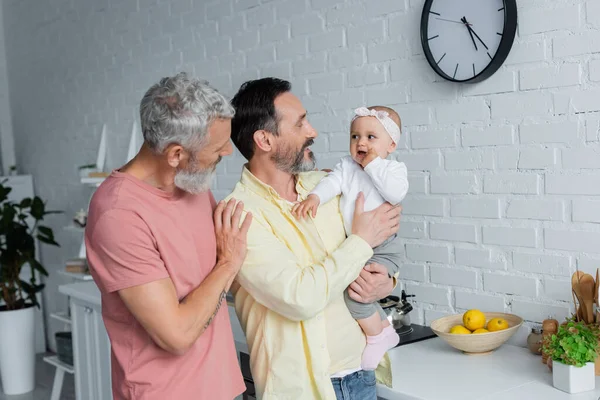 Cheerful homosexual parents holding child in kitchen — Stock Photo