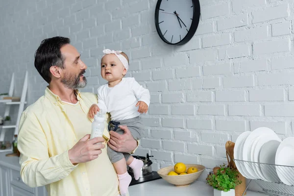 Hombre maduro sosteniendo bebé hija y biberón de leche en la cocina - foto de stock