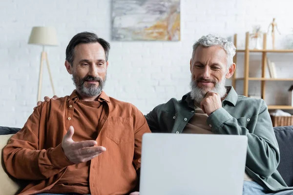 Lächelnder homosexueller Mann zeigt auf Laptop in der Nähe seines Partners zu Hause — Stockfoto