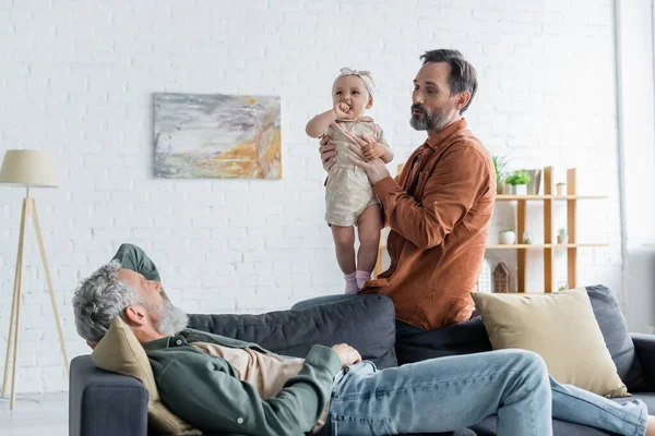 Mature homosexual man holding child near partner on couch — Stock Photo