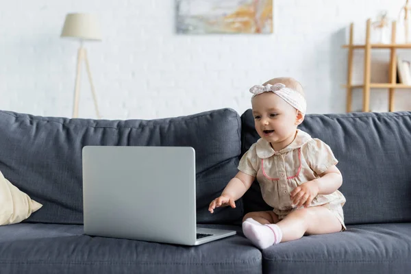 Alegre niña sentada cerca del ordenador portátil en el sofá - foto de stock