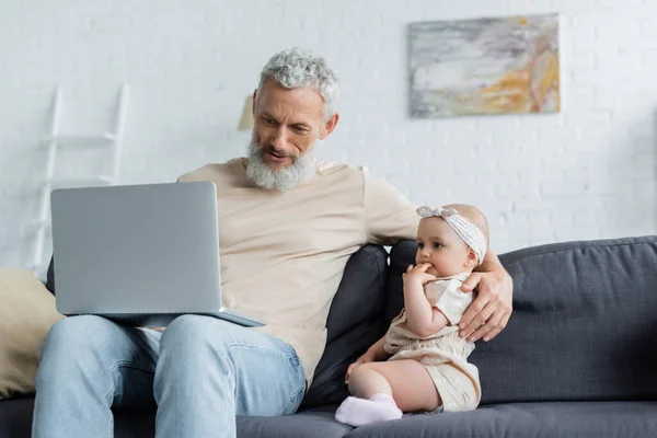 Mature man holding laptop and embracing baby girl on couch — Stock Photo