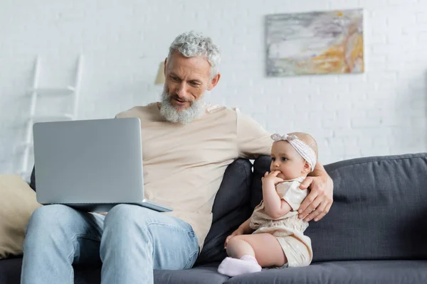 Uomo maturo che tiene il computer portatile e abbraccia la bambina sul divano — Foto stock