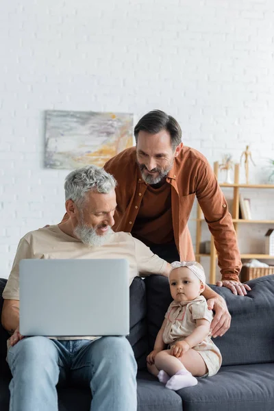 Smiling homosexual man standing near partner with laptop hugging daughter on couch — Stock Photo