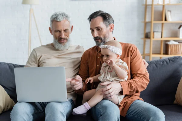 Homossexual homem apontando para laptop perto de bebê filha e parceiro em casa — Fotografia de Stock