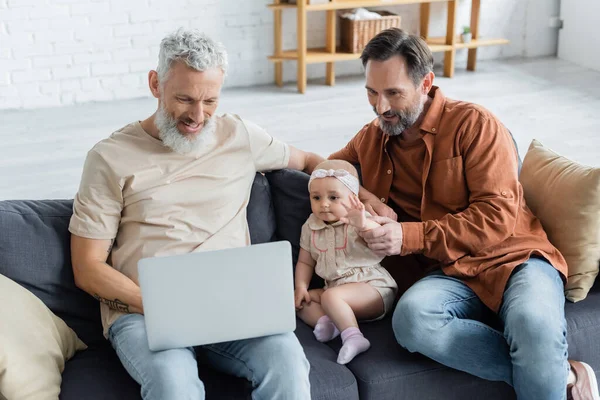 Sonriendo pareja del mismo sexo usando el ordenador portátil cerca de la hija bebé en el sofá - foto de stock