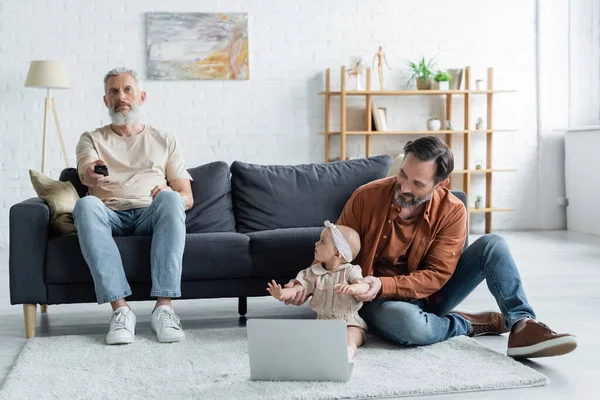 Homosexual parent sitting near daughter and laptop while partner watching tv — Stock Photo