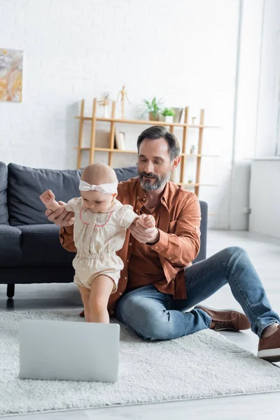 Sonriente hombre cogido de la mano de su hija pequeña cerca de la computadora portátil en la alfombra - foto de stock