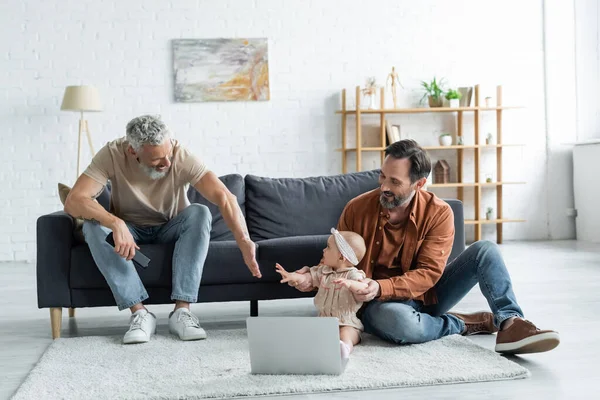 Sonriente pareja homosexual jugando con la hija del bebé cerca de la computadora portátil en la alfombra en casa - foto de stock