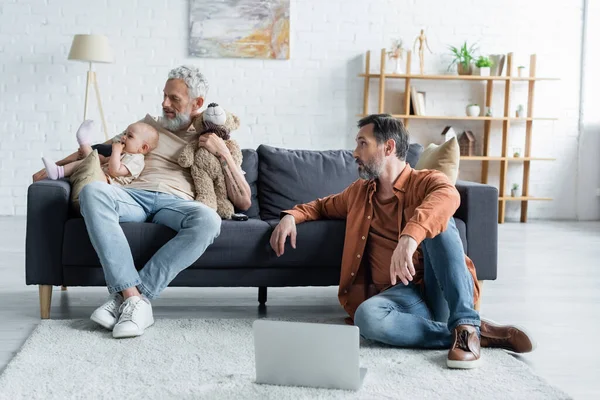 Homosexual family with soft toy and laptop playing with baby daughter at home — Stock Photo