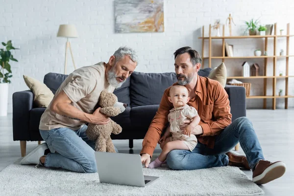 Same sex family with baby daughter using laptop and holding soft toy — Stock Photo