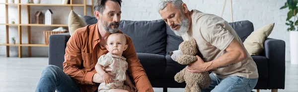 Гомосексуалист держит мягкую игрушку рядом с партнером с дочерью в гостиной, баннер — стоковое фото
