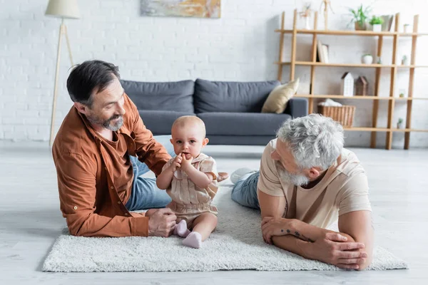 Smiling homosexual couple lying near toddler daughter on carpet — Stock Photo